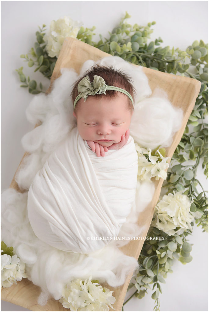 10 day old newborn baby girl photographed swaddled in white laying on white fluff with greenery