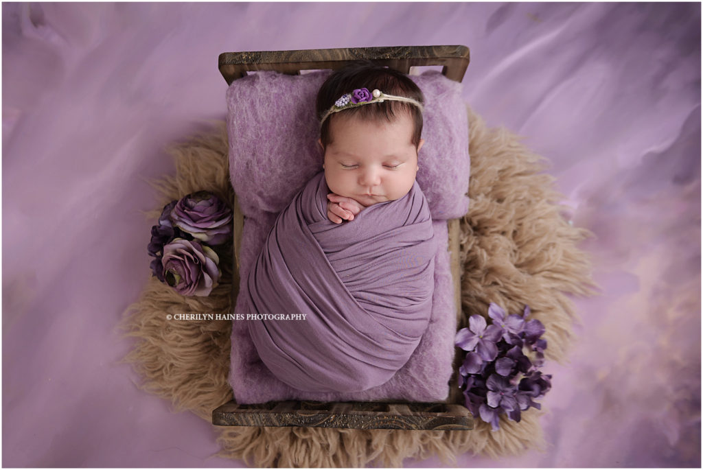 3 week old newborn baby girl photographed swaddled in purple laying on tiny wooden bed  laying on a purple backdrop; photographed by cherilyn Haines photography in baton rouge, louisiana