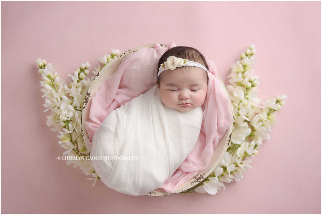 2 month old newborn baby girl photographed swaddled in white on pink backdrop with white flowers
