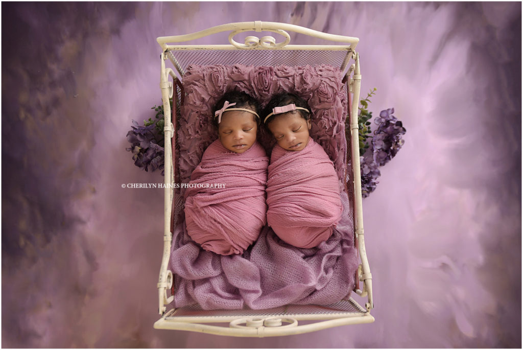 two week old newborn twin girls photographed by cherilyn haines photography in new Orleans louisiana; babies swaddled in pink laying in antique iron bed on purple backdrop