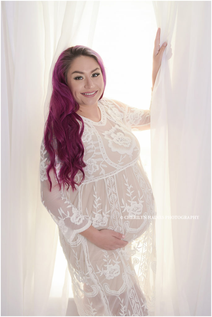 sneak peek of maternity portrait session in Baton Rouge, Louisiana; pregnant mom wearing white lace dress with purple hair, standing in front of bright, white curtains