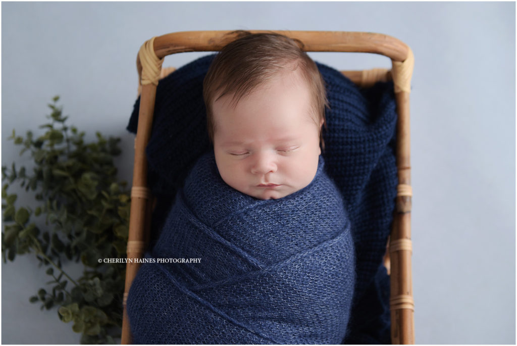 2 week old sleeping newborn baby boy photographed swaddled in a navy blanket laying in a wicker bed with greenery; photographed by cherilyn Haines photography in baton rouge, louisiana 
