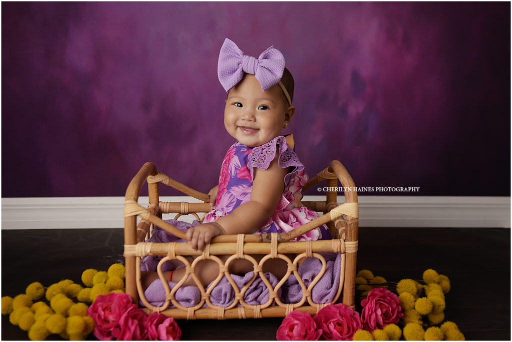 fine art baby portraits in baton rouge, louisiana with cherilyn haines photography; 6 month old baby girl posed in basket with yellow and pink flowers on a purple backdrop