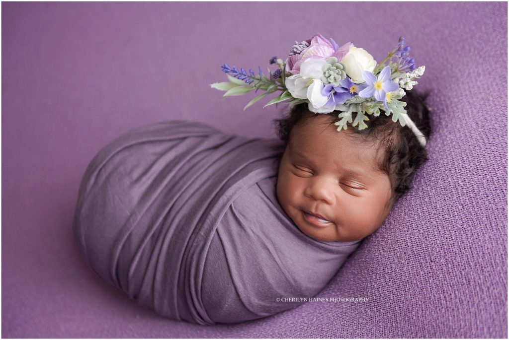 smiling newborn baby girl swaddled in purple on purple blanket with large floral headband