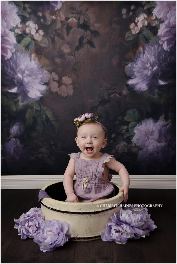 This sweet, happy 6 month old baby is a member of Cherilyn Haines Photography's baby plan and came to the studio for her sitter portrait session. She was photographed in the Baton Rouge, Louisiana location. The background chosen to match her purple romper was a dark flower backdrop with purple and lavender flowers. She was also photographed sitting in a bucket surrounded by coordinating flowers. 