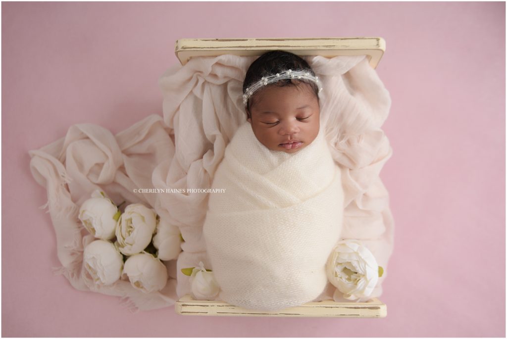 newborn baby girl photographed sleeping in a tiny antique white bed swaddled in white with white peonies on a pink backdrop; cherilyn haines photography