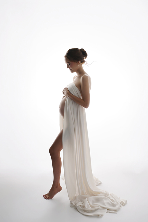 Artistic backlit maternity portrait of a pregnant woman draped in white satin fabric highlighting her baby bump in a Baton Rouge studio.