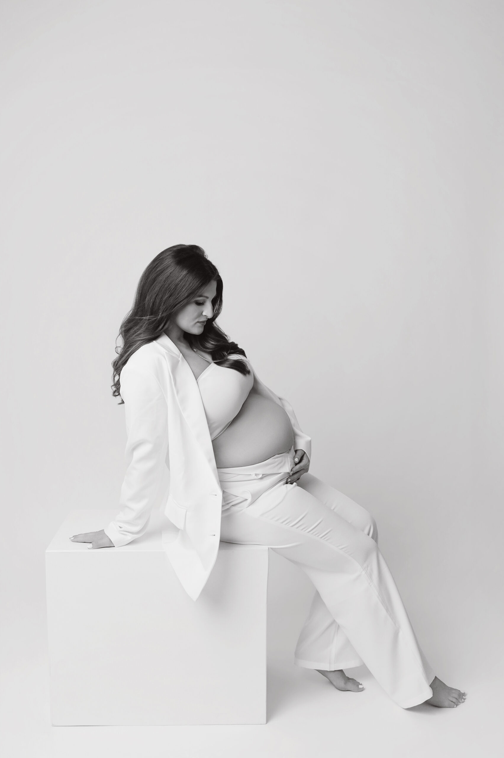 Black and white editorial maternity portrait of pregnant woman in white suit sitting on white box in Baton Rouge photography studio