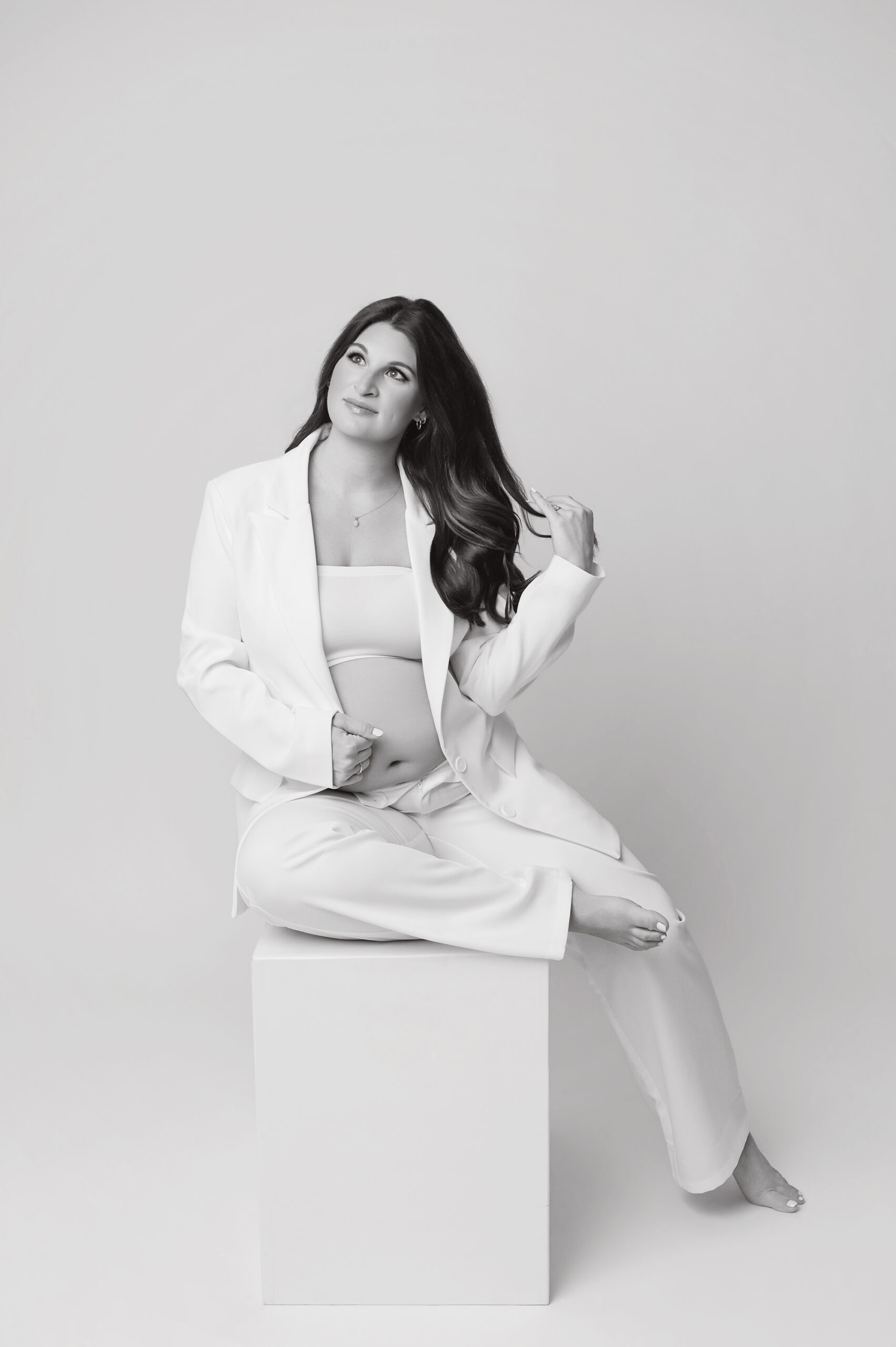 Pregnant mother in white suit sitting on white box playing with hair during black and white maternity session in Baton Rouge