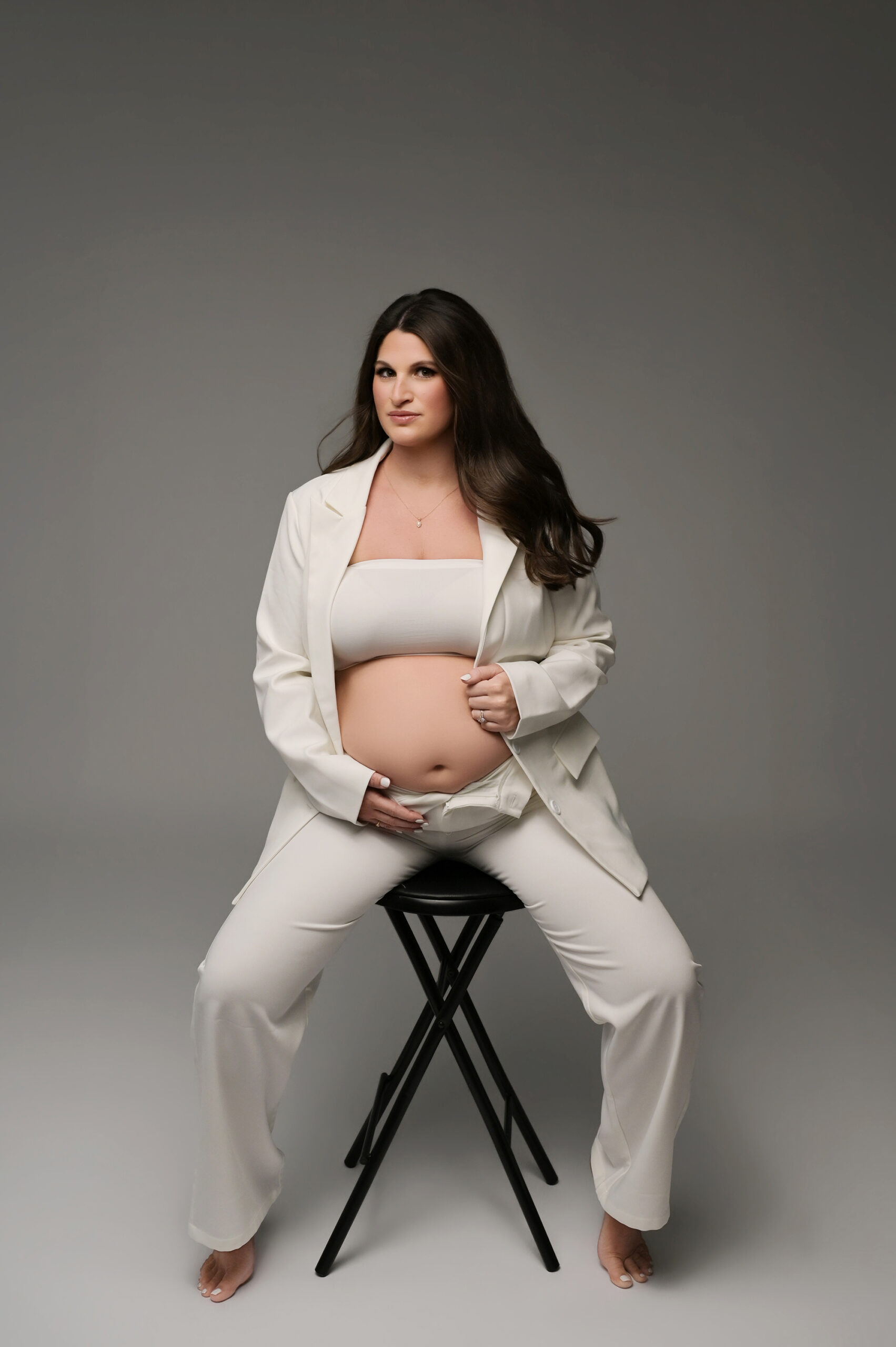 Dramatic maternity portrait of pregnant woman in white suit sitting on black stool with studio lighting