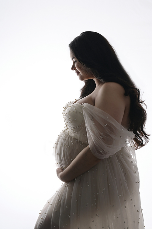 Ethereal backlit maternity portrait of an expectant mother wearing a white gown with pearl accents in a Baton Rouge photography studio.