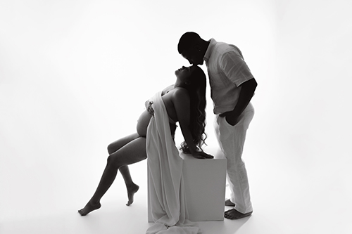 Black and white maternity portrait of an expectant father kissing mom's forehead while she sits draped in flowing white fabric.