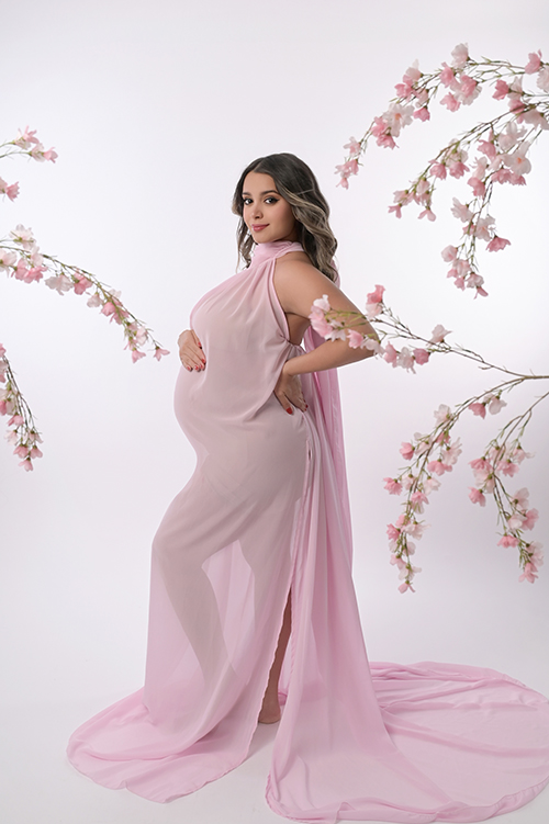 Expectant mother wearing a sheer pink maternity gown surrounded by pink florals during a luxury studio maternity session in Baton Rouge.