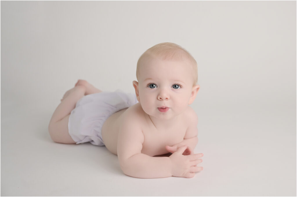 6 month old baby boy wearing a white diaper cover with his hands in his mouth on a soft white blanket backdrop