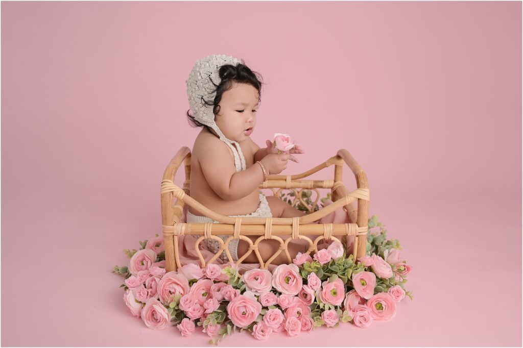 6-month-old baby girl wearing a bonnet sitting in a basket surrounded by pink roses