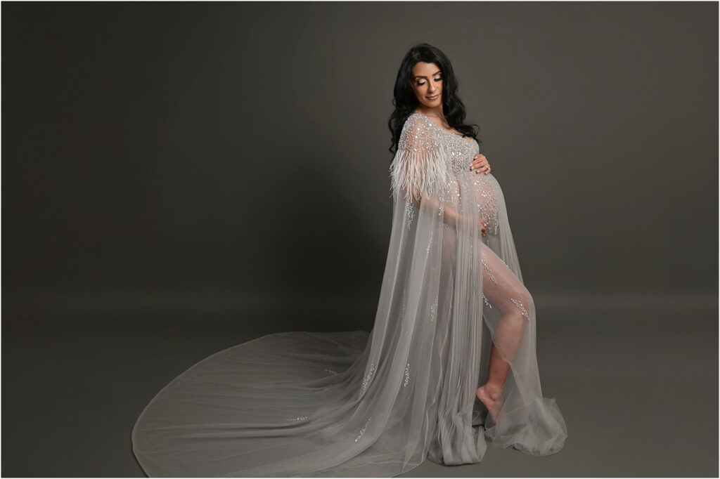 Pregnant woman wearing a silver beaded maternity gown with a flowing cape during a studio maternity portrait session