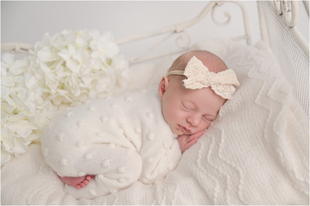 Newborn baby girl in a white footed romper lying on a white iron bed with florals