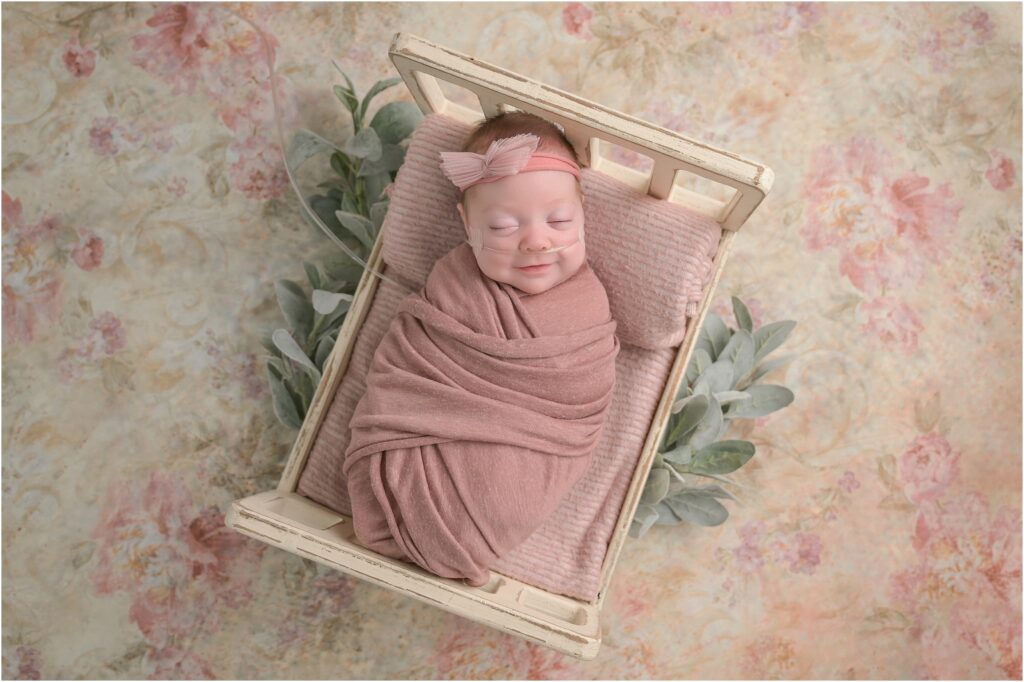 Premature newborn baby girl swaddled in pink on a tiny wooden bed with a floral backdrop smiling